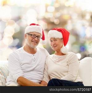 family, holidays, christmas, age and people concept - happy senior couple in santa helper hats sitting on sofa over lights background