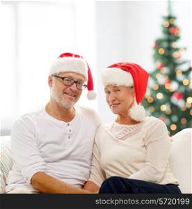 family, holidays, christmas, age and people concept - happy senior couple in santa helper hats sitting on sofa over living room and christmas tree background