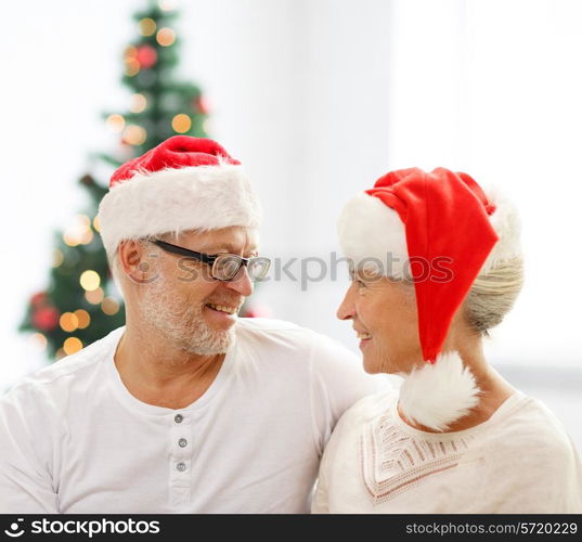 family, holidays, christmas, age and people concept - happy senior couple in santa helper hats sitting on sofa over living room and christmas tree background