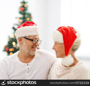 family, holidays, christmas, age and people concept - happy senior couple in santa helper hats sitting on sofa over living room and christmas tree background