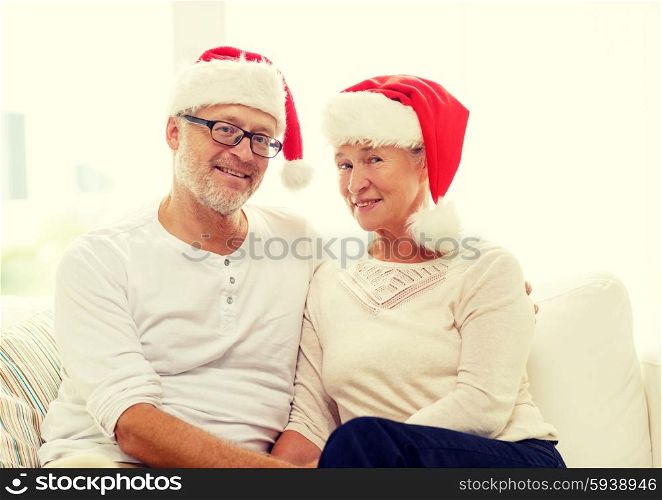 family, holidays, christmas, age and people concept - happy senior couple in santa helper hats sitting on sofa at home