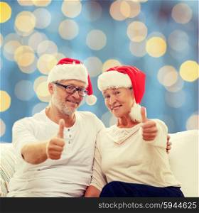 family, holidays, christmas, age and people concept - happy senior couple in santa helper hats sitting on sofa and showing thumbs up gesture over blue lights background
