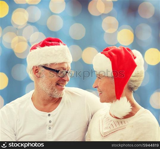 family, holidays, christmas, age and people concept - happy senior couple in santa helper hats sitting on sofa over blue lights background