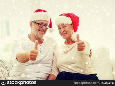 family, holidays, christmas, age and people concept - happy senior couple in santa helper hats sitting on sofa and showing thumbs up gesture at home