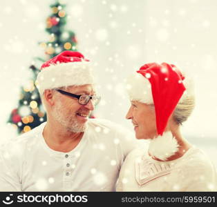 family, holidays, christmas, age and people concept - happy senior couple in santa helper hats looking to each other at home