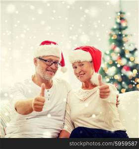 family, holidays, christmas, age and people concept - happy senior couple in santa helper hats sitting on sofa over living room and christmas tree background