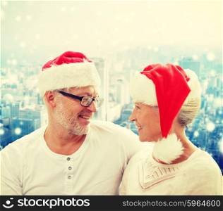 family, holidays, christmas, age and people concept - happy senior couple in santa helper hats sitting on sofa over snowy city background