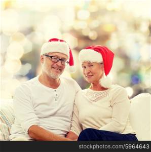 family, holidays, christmas, age and people concept - happy senior couple in santa helper hats sitting on sofa over lights background