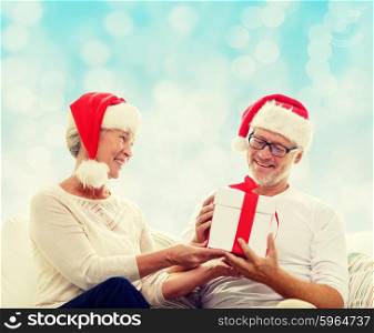 family, holidays, christmas, age and people concept - happy senior couple in santa helper hats with gift box over blue lights background