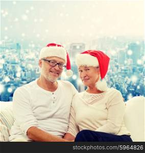 family, holidays, christmas, age and people concept - happy senior couple in santa helper hats sitting on sofa over snowy city background