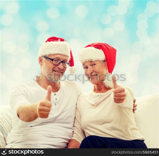 family, holidays, christmas, age and people concept - happy senior couple in santa helper hats sitting on sofa over blue lights background and showing thumbs up gesture