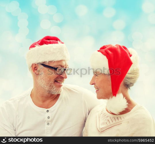 family, holidays, christmas, age and people concept - happy senior couple in santa helper hats sitting on sofa over blue lights background