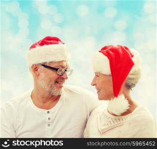 family, holidays, christmas, age and people concept - happy senior couple in santa helper hats sitting on sofa over blue lights background