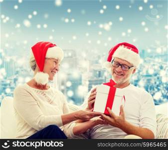 family, holidays, christmas, age and people concept - happy senior couple in santa helper hats with gift box over snowy city background
