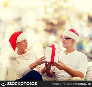 family, holidays, christmas, age and people concept - happy senior couple in santa helper hats with gift box over lights background
