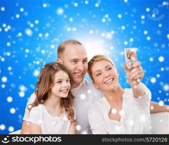 family, holidays, christmas technology and people concept - smiling mother, father and little girl making selfie with camera over blue snowy background