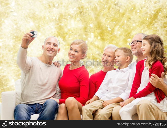 family, holidays, generation, christmas and people concept - smiling family with camera taking selfie and sitting on couch over yellow lights background