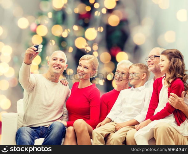 family, holidays, generation, christmas and people concept - smiling family with camera taking selfie and sitting on couch over tree lights background