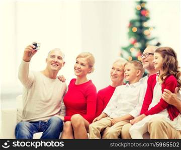 family, holidays, generation, home and people concept - smiling family with camera taking selfie and sitting on couch over living room with christmas tree background