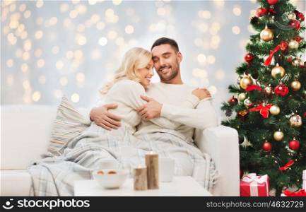 family, holidays, love and people concept - happy couple covered with plaid hugging on sofa over christmas tree and lights background