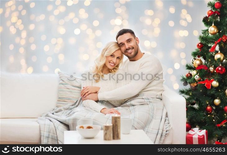 family, holidays, love and people concept - happy couple covered with plaid sitting on sofa over christmas tree and lights background