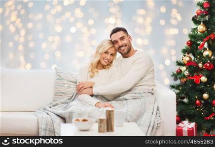 family, holidays, love and people concept - happy couple covered with plaid sitting on sofa over christmas tree and lights background