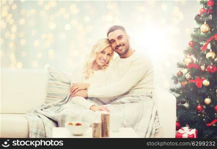 family, holidays, love and people concept - happy couple covered with plaid sitting on sofa over christmas tree and lights background. happy couple hugging over christmas tree