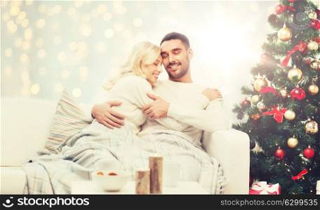 family, holidays, love and people concept - happy couple covered with plaid hugging on sofa over christmas tree and lights background. happy couple hugging over christmas tree