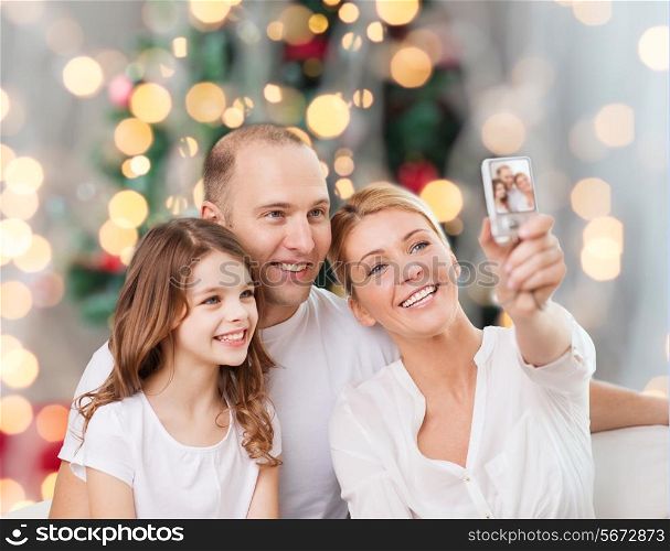 family, holidays, technology and people concept - smiling mother, father and little girl making selfie with camera over christmas tree lights background