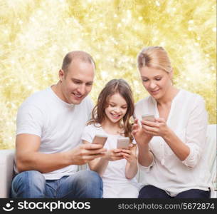 family, holidays, technology and people concept - smiling mother, father and little girl with smartphones over yellow lights background