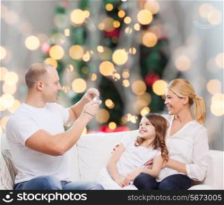 family, holidays, technology and people concept - smiling mother, father and little girl with camera over christmas tree lights background