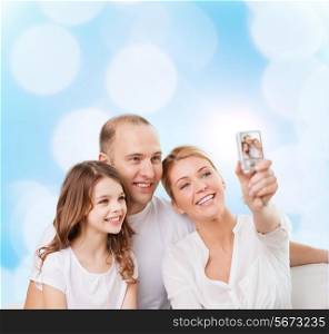 family, holidays, technology and people concept - smiling mother, father and little girl making selfie with camera over blue lights background