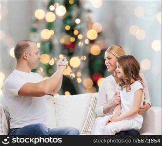 family, holidays, technology and people concept - smiling mother, father and little girl with camera over christmas tree lights background