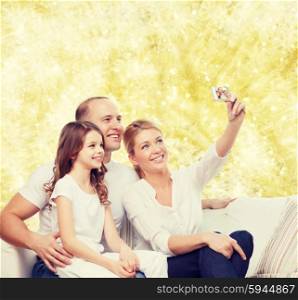 family, holidays, technology and people concept - smiling mother, father and little girl making selfie with camera over yellow lights background