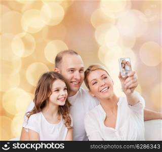 family, holidays, technology and people concept - smiling mother, father and little girl making selfie with camera over beige lights background