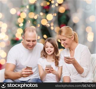family, holidays, technology and people - smiling mother, father and little girl with smartphones over christmas tree lights background