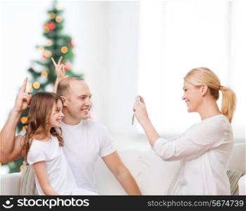 family, holidays, technology and people - smiling mother, father and little girl with camera over living room and christmas tree background