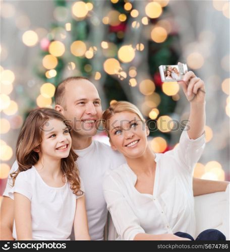family, holidays, technology and people - smiling mother, father and little girl making selfie with camera over christmas tree lights background