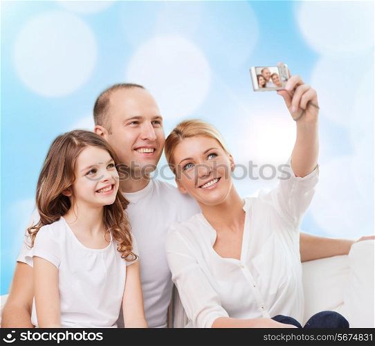 family, holidays, technology and people - smiling mother, father and little girl making selfie with camera over blue lights background