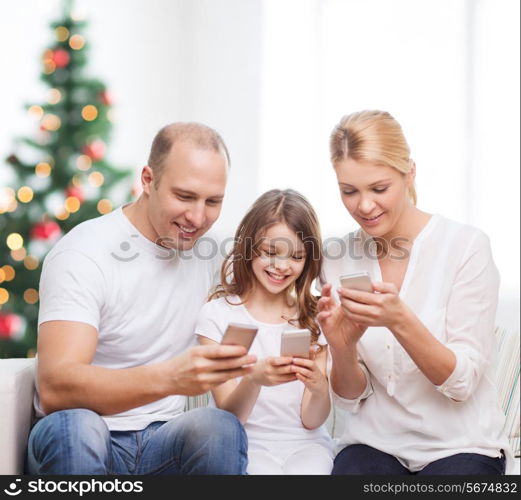 family, holidays, technology and people - smiling mother, father and little girl with smartphones over living room and christmas tree background