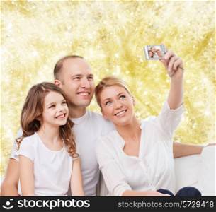 family, holidays, technology and people - smiling mother, father and little girl making selfie with camera over yellow lights background