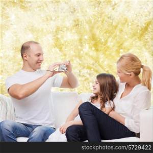family, holidays, technology and people - smiling mother, father and little girl with camera over yellow lights background