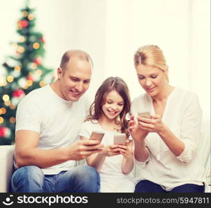 family, holidays, technology and people - smiling mother, father and little girl with smartphones over living room and christmas tree background