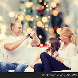 family, holidays, technology and people - smiling mother, father and little girl with camera over christmas tree lights background