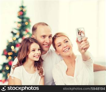family, holidays, technology and people - smiling mother, father and little girl making selfie with camera over living room and christmas tree background