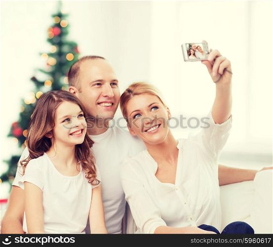 family, holidays, technology and people - smiling mother, father and little girl making selfie with camera over living room and christmas tree background