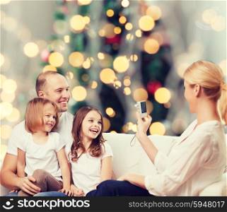 family, holidays, technology and people - smiling mother, father and little girls with camera over christmas tree lights background