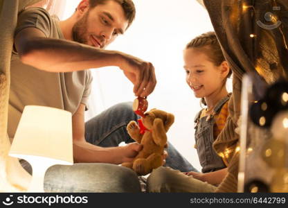 family, hygge and people concept - happy father and little daughter playing with teddy bear and toy tea pot in kids tent at night at home. happy family playing with toy in kids tent at home