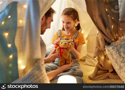 family, hygge and people concept - happy father with teddy bear toy and little daughter playing in kids tent at night at home. happy family playing with toy in kids tent at home