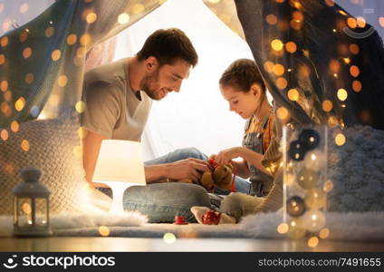 family, hygge and people concept - happy father with teddy bear toy and little daughter playing in kids tent at night at home. happy family playing with toy in kids tent at home